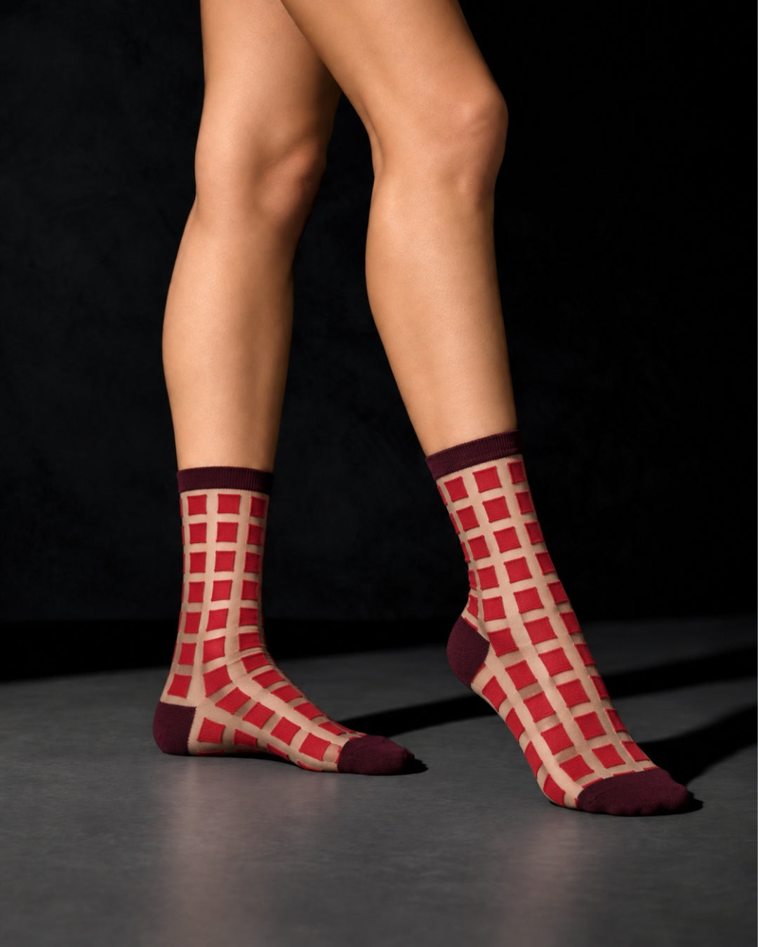 Model wearing sheer Ginza Cerise socks with a red grid pattern and burgundy cuffs, heels and toes, standing on a gray floor against a black background