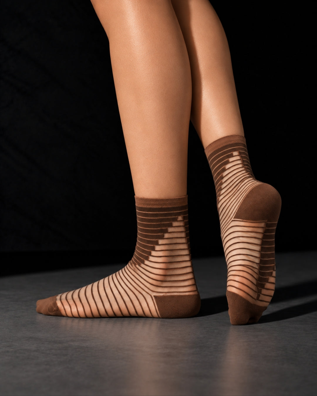 Model wearing sheer Shibuya Moka socks with brown and caramel stripes, one heel lifted to show the back of the sock, against a black background