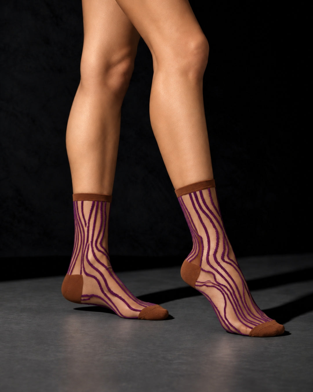 Model wearing sheer Superkilen Prune socks with wavy purple lines and brown cuffs, heels and toes, standing on a gray floor against a black background