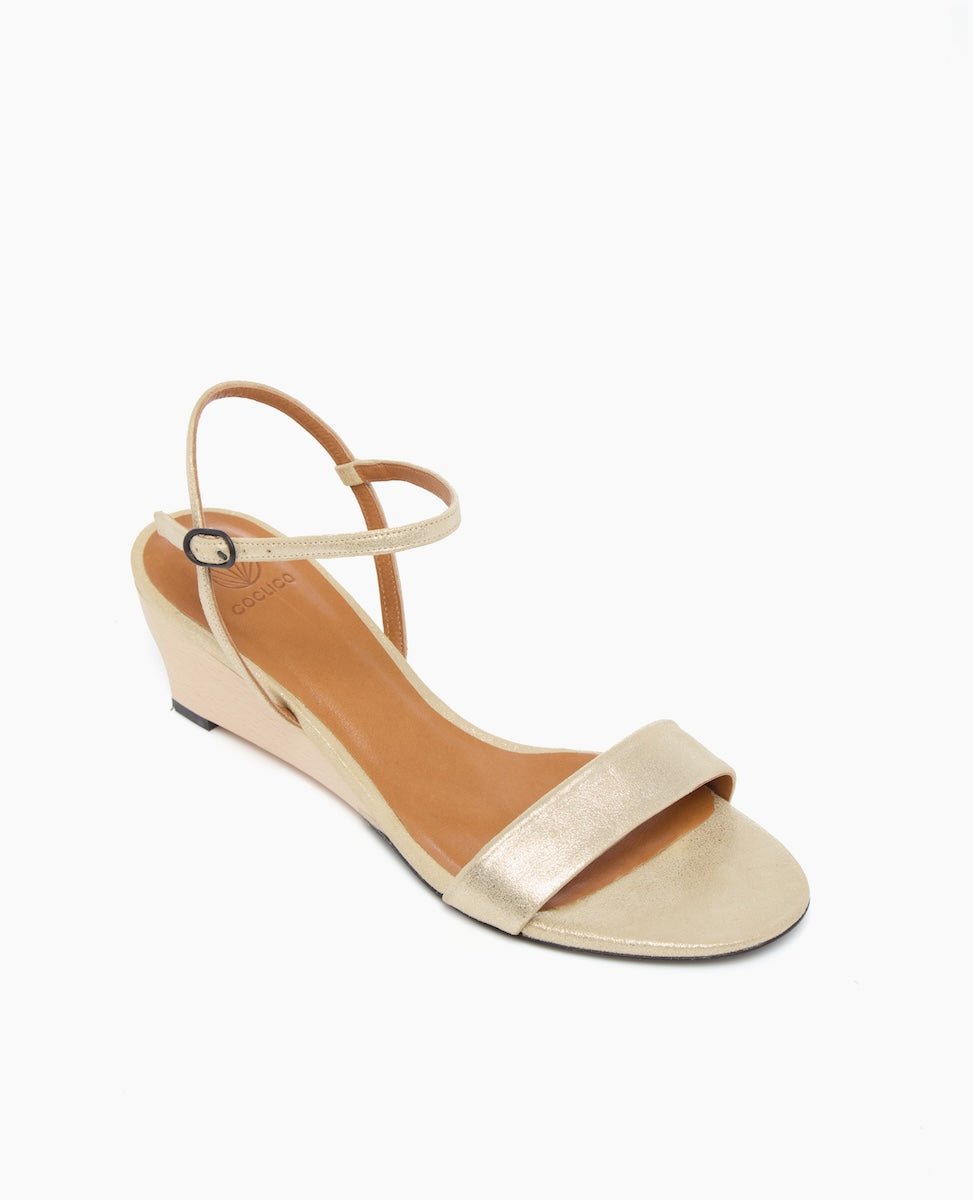A sleek, open-toe sandal with a metallic gold finish, featuring a low wedge heel, a single front strap, and a thin ankle strap with a small buckle.