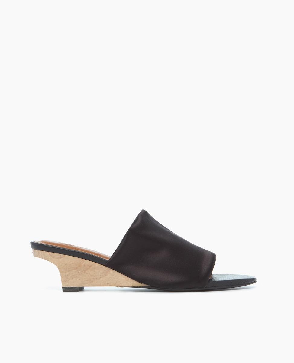 A sleek, black leather slide sandal with a smooth texture, featuring a minimalist design and a light wooden wedge heel.