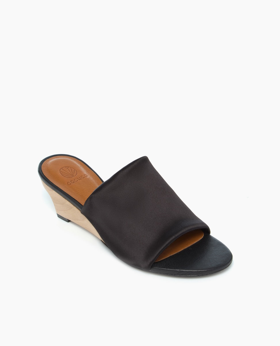 A sleek black open-toe mule with a smooth satin-like upper, featuring a low wedge heel in a light wood finish and a tan insole with a subtle logo imprint.