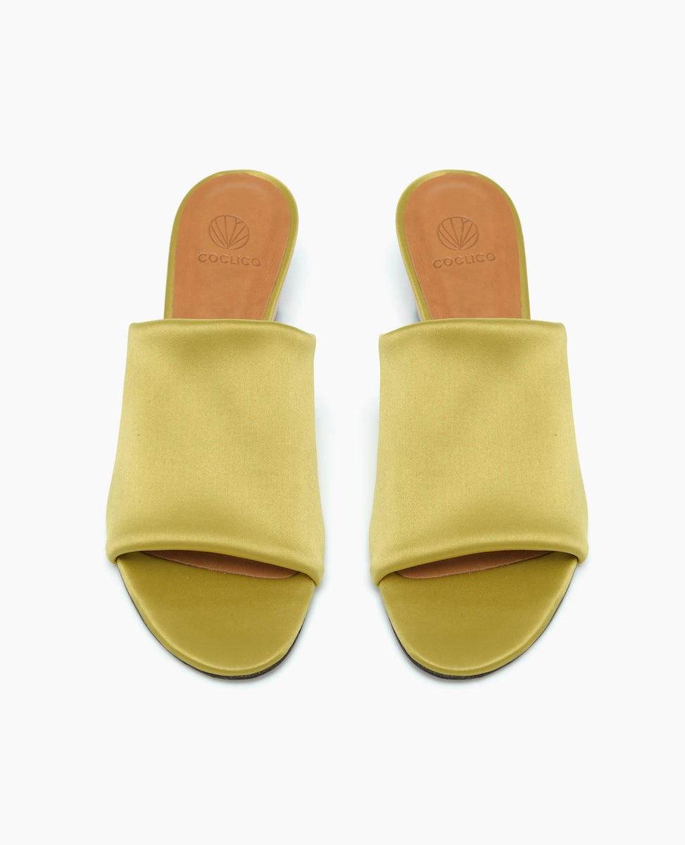 A pair of open-toed, slip-on sandals with a mustard yellow fabric upper, tan leather insole, and a flat sole, featuring a minimalist design with a smooth texture and subtle branding on the insole.