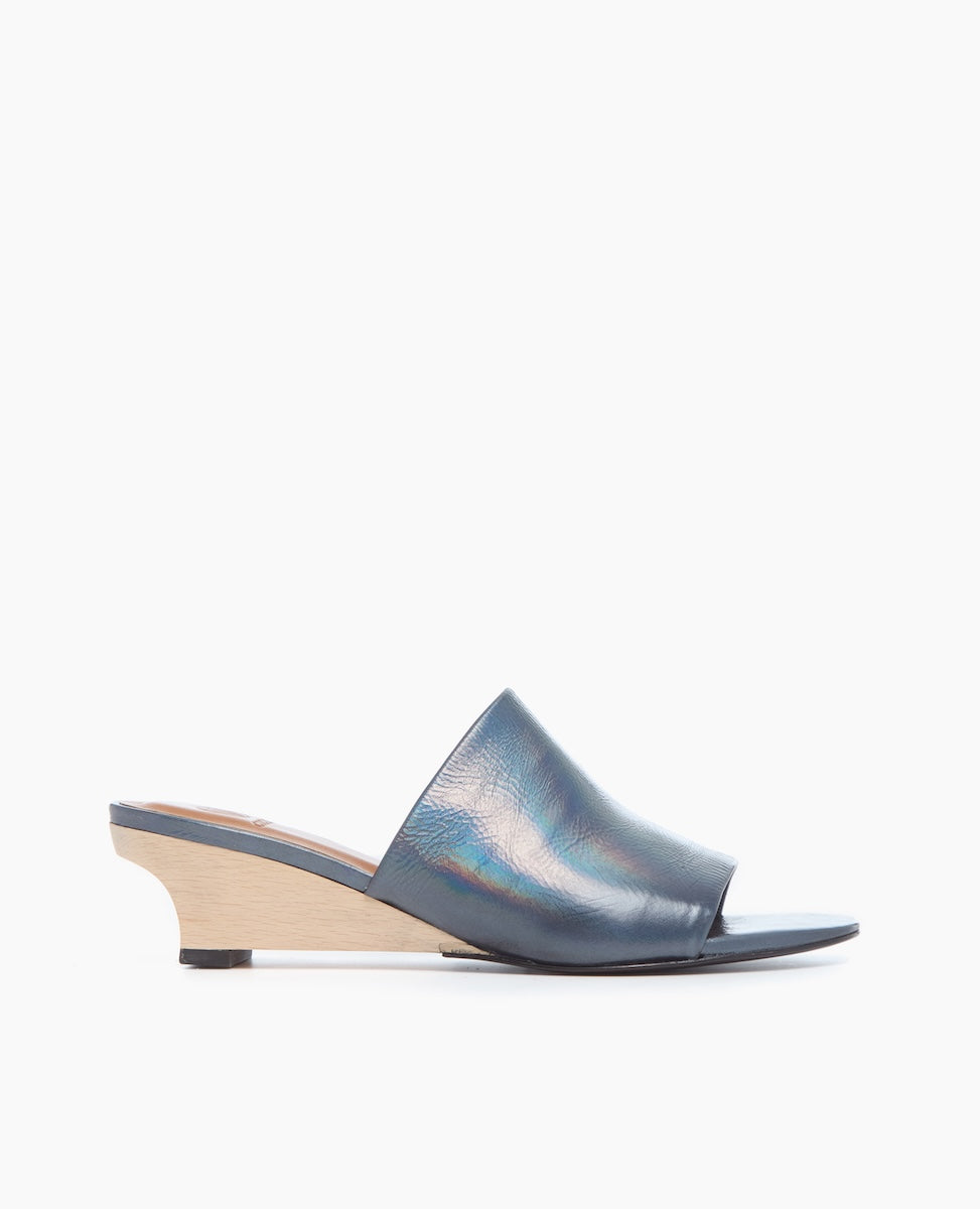 A sleek, open-toe mule sandal featuring a smooth, iridescent blue leather upper, a low wooden wedge heel, and a minimalist design with a single wide strap across the foot.