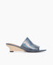 A sleek, open-toe mule sandal featuring a smooth, iridescent blue leather upper, a low wooden wedge heel, and a minimalist design with a single wide strap across the foot. 2