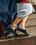 A person wearing a light gray skirt and dark blue shoes sits on a red chair, with sunlight casting shadows on the speckled floor. 7