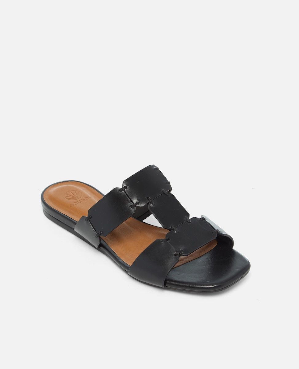 Angled view of a black vachetta leather T-strap slide sandal with octagonal hand stitched panels, square toe, and flat sole, on a white background.