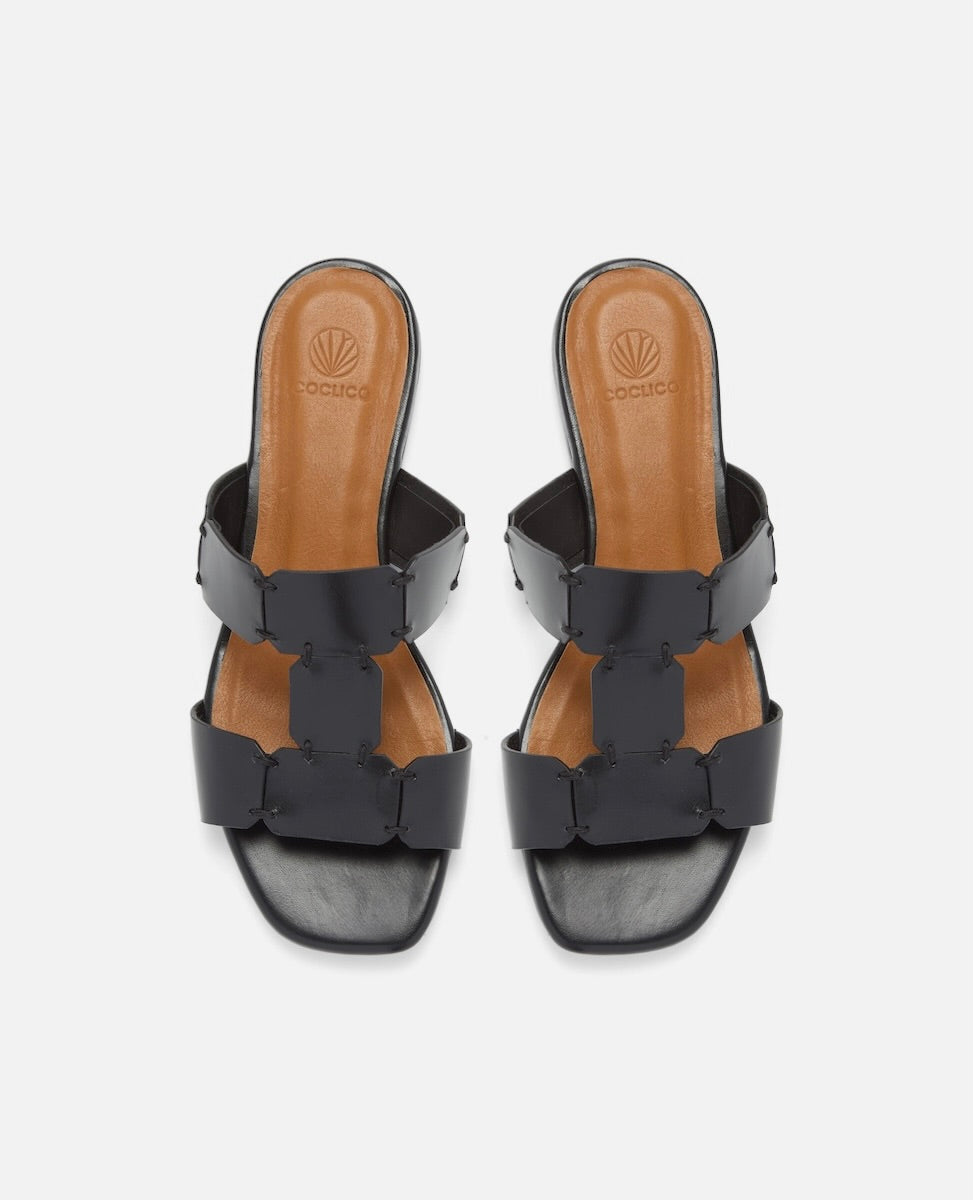 Top view of a black vachetta leather T-strap slide sandal with octagonal hand stitched panels, square toe, and flat sole, on a white background.