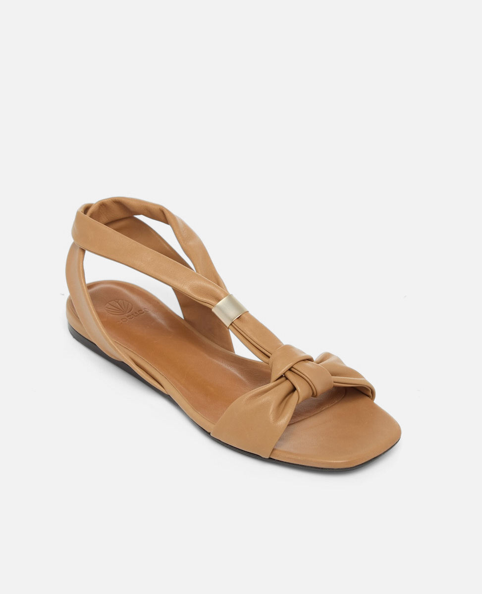 Angled view of a tan leather flat sandal with tubular straps, an adjustable brushed-metal keeper at the center strap, toe knot detail, and a square toe, on a white background.