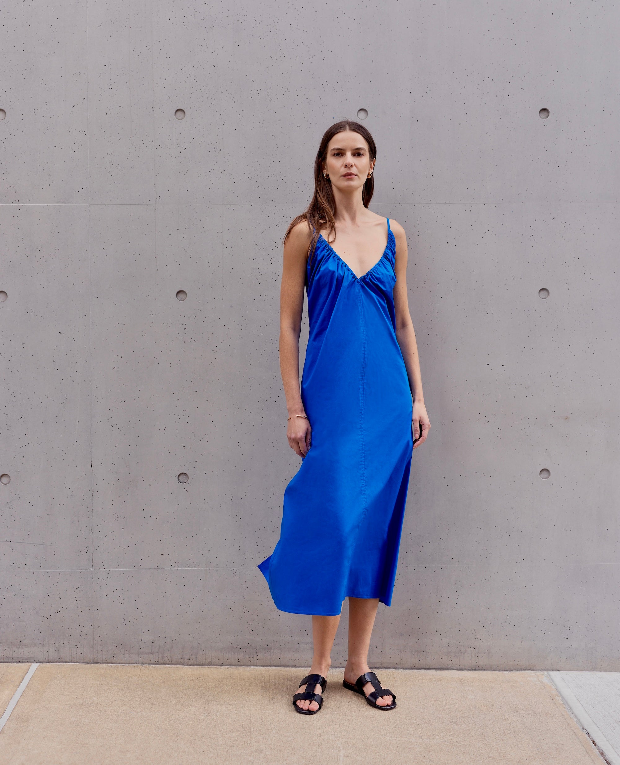 Woman wearing a blue dress and black sandals standing against a gray concrete wall.