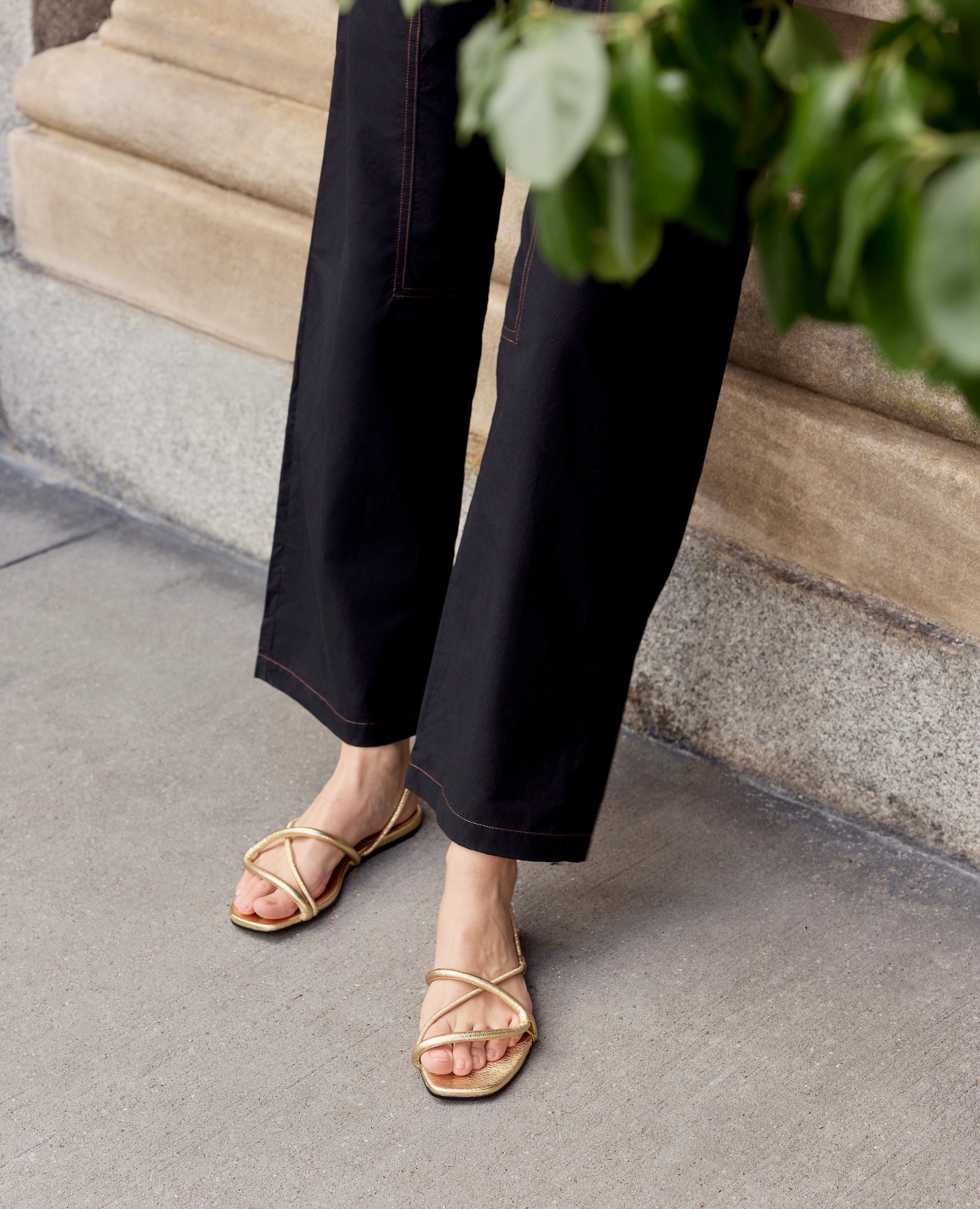 Person wearing black pants and gold sandals on a sidewalk.