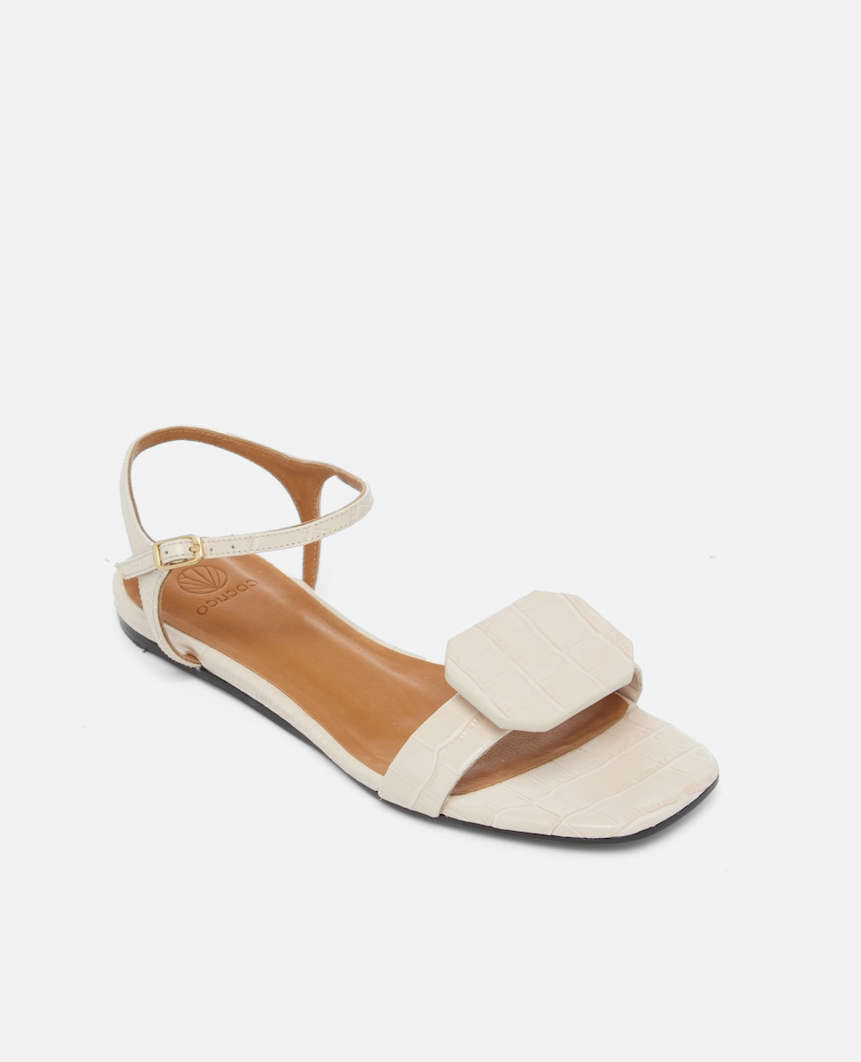 Angled view of a bone-colored croc-embossed leather flat sandal with a slim ankle strap and a octagonal accent on the front strap, square toe, on a white background.