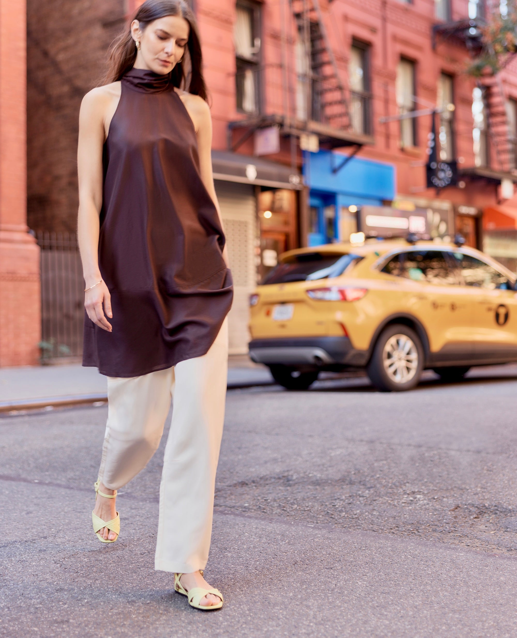 Woman in a brown sleeveless top and light pants walking on a city street with a yellow taxi in the background.