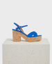 A vibrant blue sandal with a cork block heel, featuring crisscross straps and an adjustable ankle buckle, resting on a textured white platform. 1