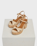 Beige strappy sandals with a cork block heel, featuring a crisscross design on the front and an adjustable ankle strap, resting on a textured white platform. 1