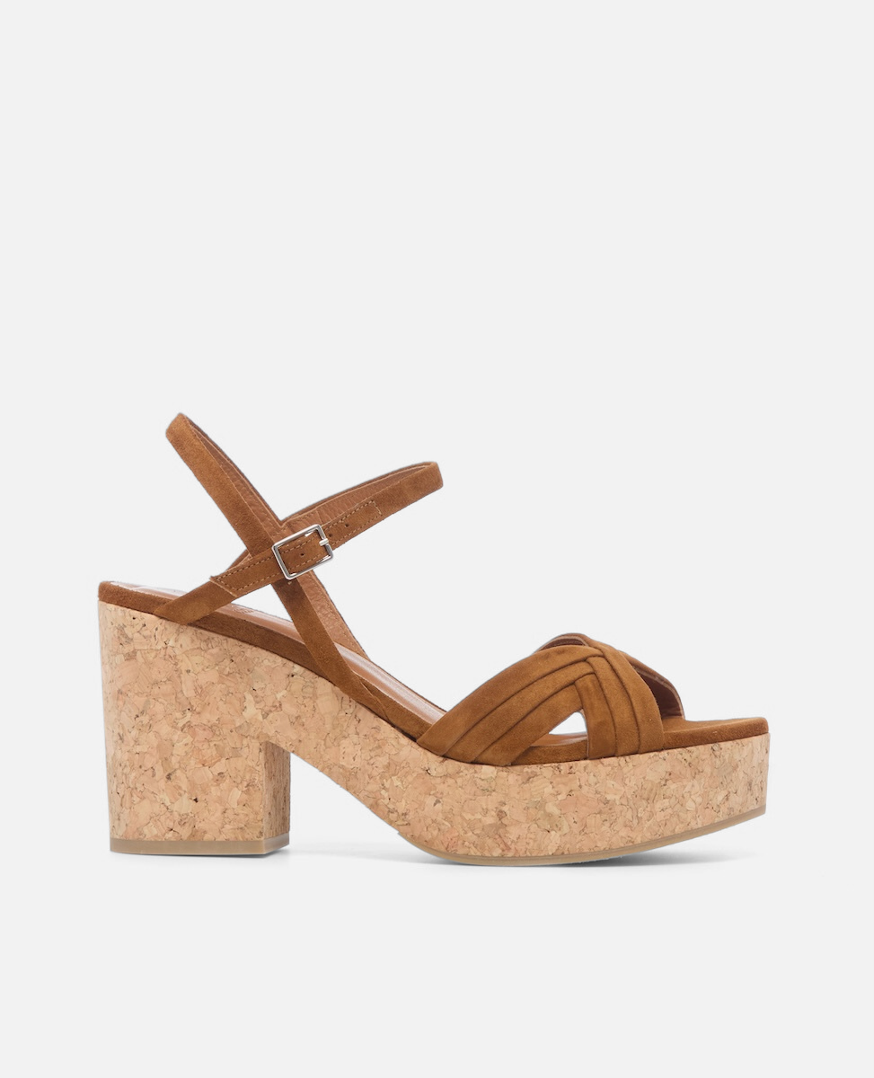 A brown suede sandal with a cork block heel, featuring a crisscross strap design and an adjustable ankle strap.