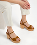 Brown suede sandals with a cork platform heel, featuring crisscross straps and an adjustable ankle buckle, worn by a person in white pants. 2