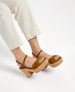 Brown suede platform sandals with cork wedge heels, featuring an open toe design and adjustable ankle straps, worn by a person in light-colored pants. 3