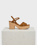 A brown sandal with a cork block heel and platform, featuring a strappy suede upper and an adjustable ankle strap, displayed on a textured white pedestal. 1