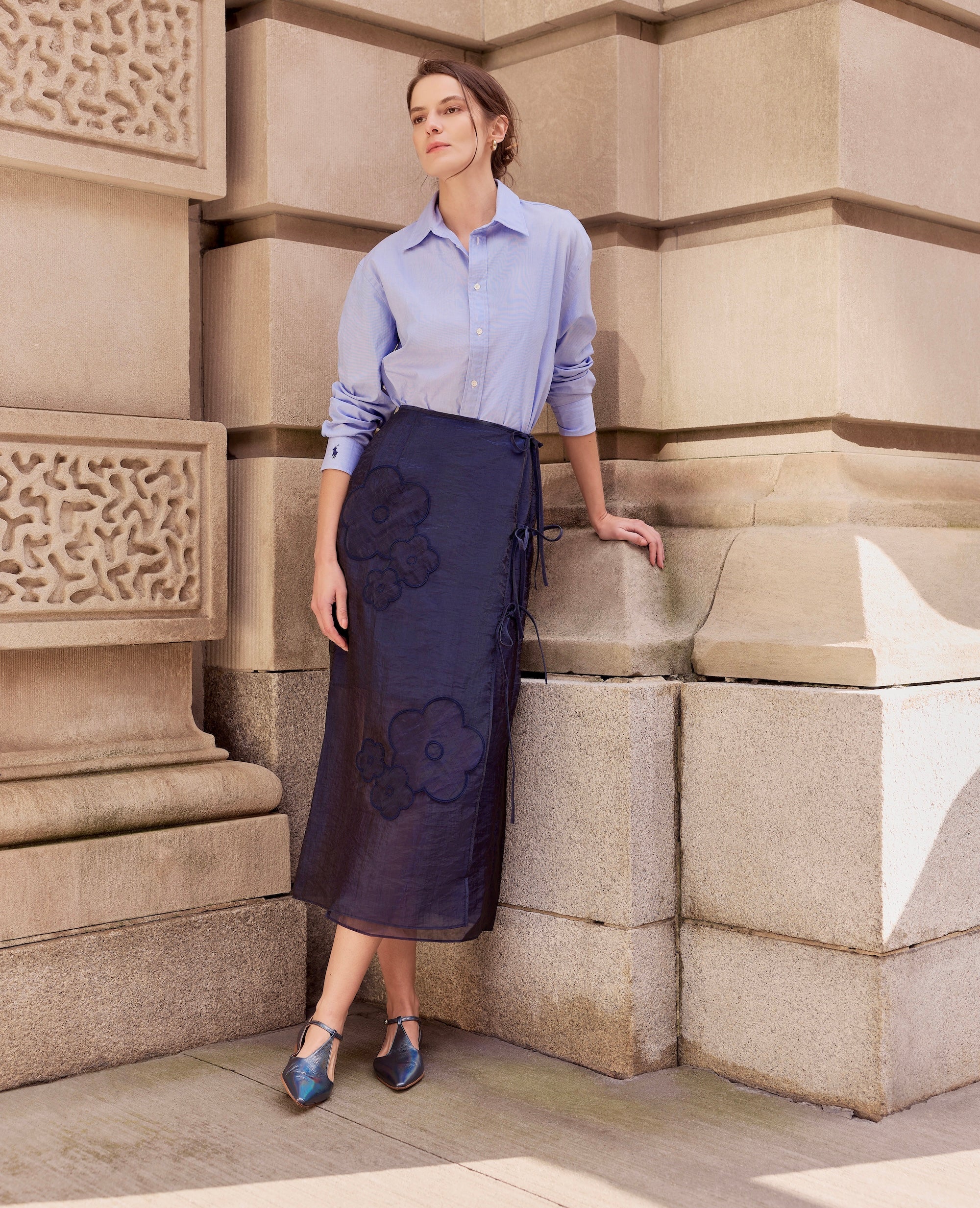 Woman in a light blue shirt, dark blue floral skirt, and iridescent shoes standing against a stone wall.