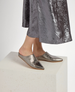 A pair of metallic silver, pointed-toe mules with a sleek, shiny texture and a flat sole, worn by a person standing on a light-colored textured block, partially covered by a shimmering silver skirt. 4