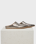 A pair of sleek, metallic silver slip-on mules with a pointed toe, flat sole, and textured leather finish, resting on a light-colored stone block. 1