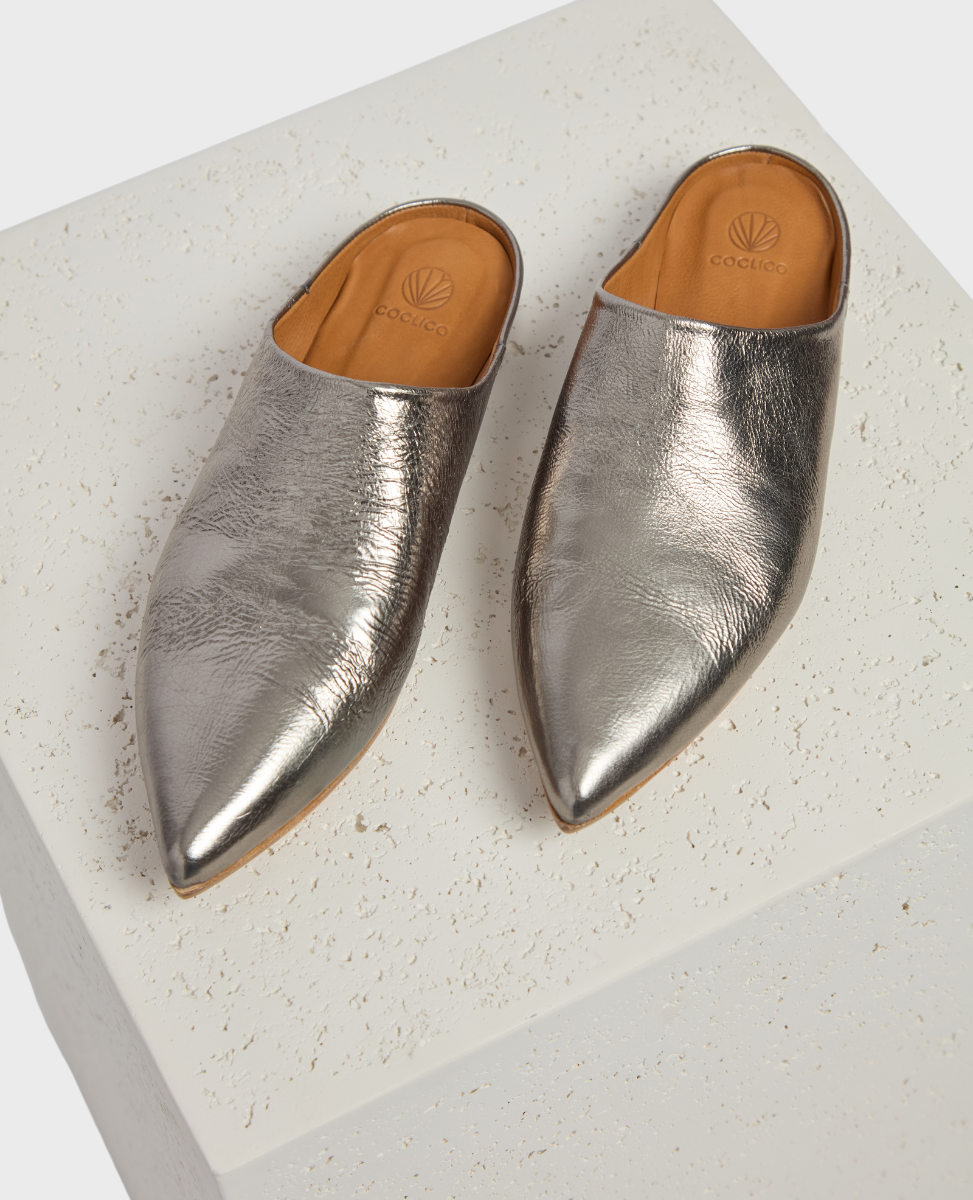 A pair of sleek, metallic silver mules with a pointed toe design, featuring a smooth, shiny leather texture and a tan leather insole with a subtle logo imprint.