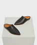 A pair of sleek, black leather mules with a pointed toe design, smooth texture, and tan interior lining, resting on a textured white cube. 1