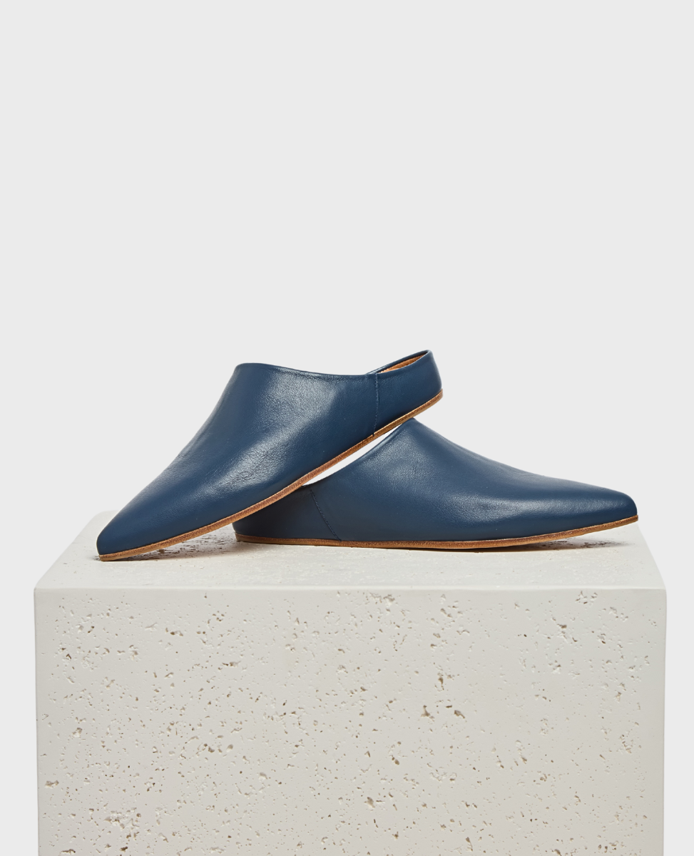 A pair of sleek, blue leather slip-on shoes with a pointed toe, flat sole, and minimalistic design, displayed on a textured, light-colored stone pedestal.