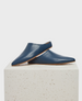 A pair of sleek, blue leather slip-on shoes with a pointed toe, flat sole, and minimalistic design, displayed on a textured, light-colored stone pedestal. 1