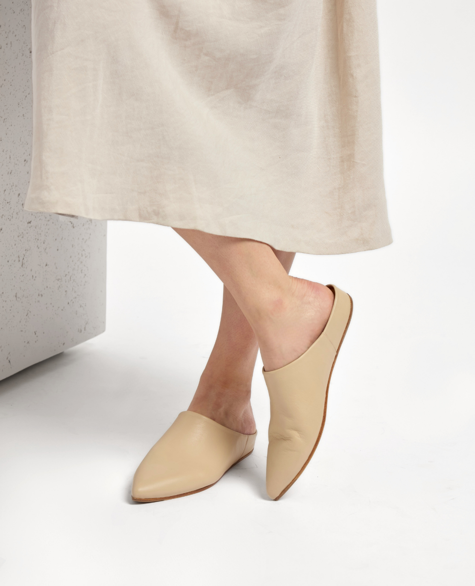 A pair of sleek, beige slip-on mules with a pointed toe, smooth leather texture, and a flat sole, worn by a person standing on a white surface.