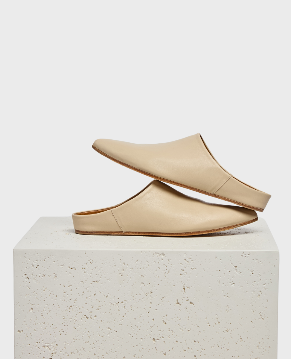 A pair of sleek, beige leather slip-on mules with a smooth texture, minimalist design, and flat soles, displayed on a textured white pedestal.