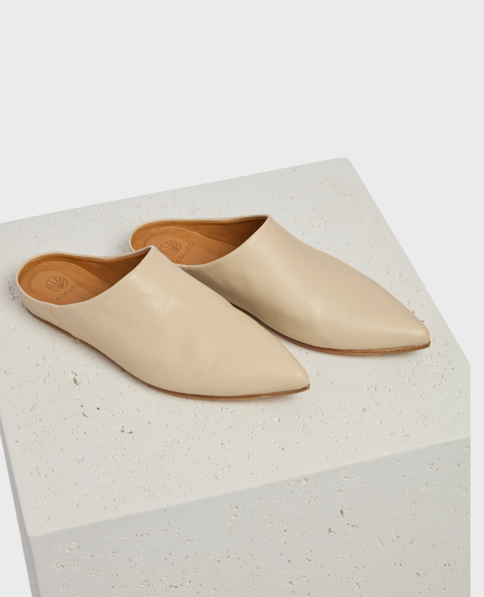 A pair of sleek, pointed-toe mules in a smooth, beige leather with a minimalist design and a flat sole, resting on a textured white surface.