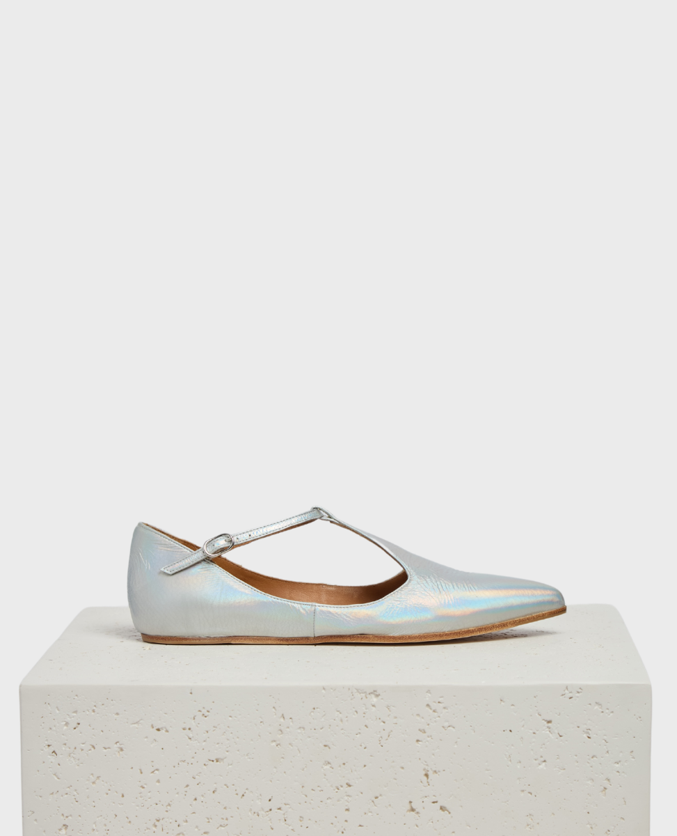 A sleek, iridescent silver flat shoe with a pointed toe, featuring a thin T-strap and a smooth, glossy texture, resting on a light-colored, textured block.