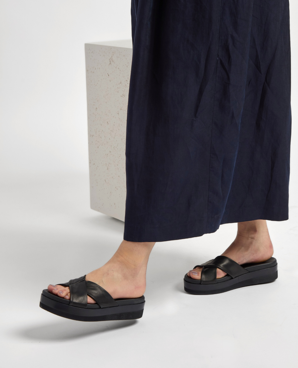 A pair of black, open-toed sandals with wide, crisscross leather straps and a thick, flat platform sole, worn by a person in a long, dark garment.