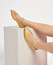 Golden, pointed-toe flats with a shiny, iridescent finish, featuring a slender ankle strap with a small buckle, resting on a textured, light-colored stone block. 2