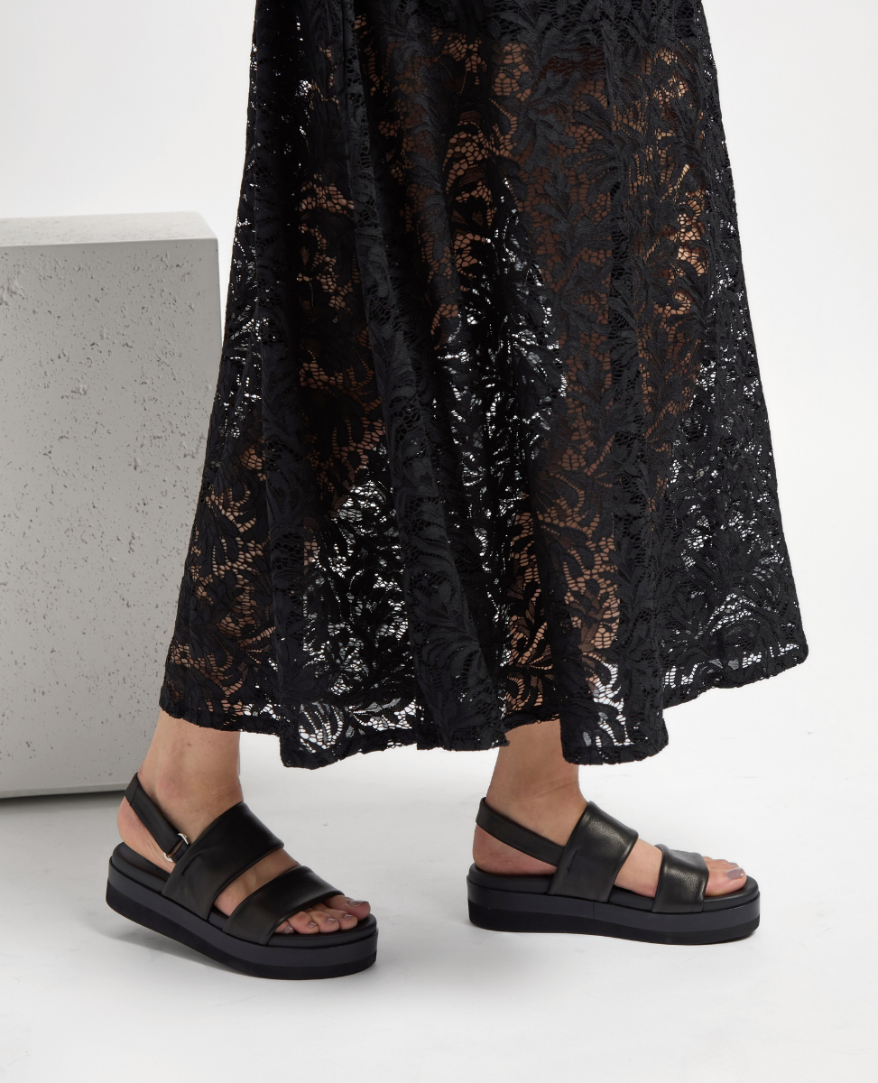 Black leather sandals with thick straps and a platform sole, featuring a matte finish and a buckle closure, worn with a long, sheer black lace skirt.