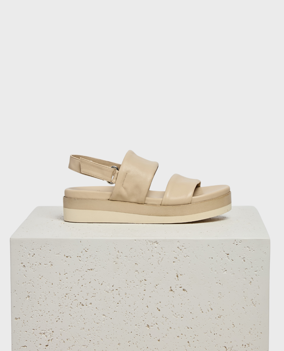 A beige sandal with a thick, cushioned sole, featuring two wide, padded straps across the foot and an adjustable slingback strap, all made from smooth leather.