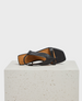 A black leather sandal with a square toe, featuring a minimalist design and a thin ankle strap, displayed on a textured white block. 1