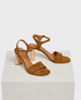 A pair of brown suede sandals with a block heel, featuring an open toe design, ankle strap with a buckle, and a smooth leather insole, resting on a textured white platform. 1