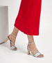 Silver strappy sandals with a holographic sheen, featuring a chunky wooden heel and a delicate ankle strap, paired with a vibrant red pleated skirt. 3