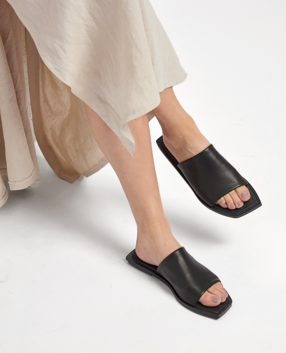 A pair of black leather slide sandals with a square toe design and flat sole, worn by a person in a flowing beige skirt.