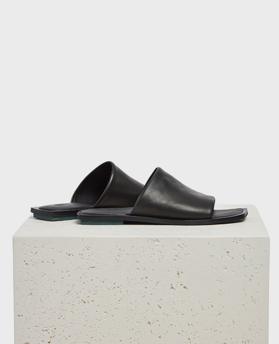 A pair of sleek, black leather slide sandals with a minimalist design, featuring a smooth texture, open toe, and flat sole, displayed on a textured, light-colored stone surface.