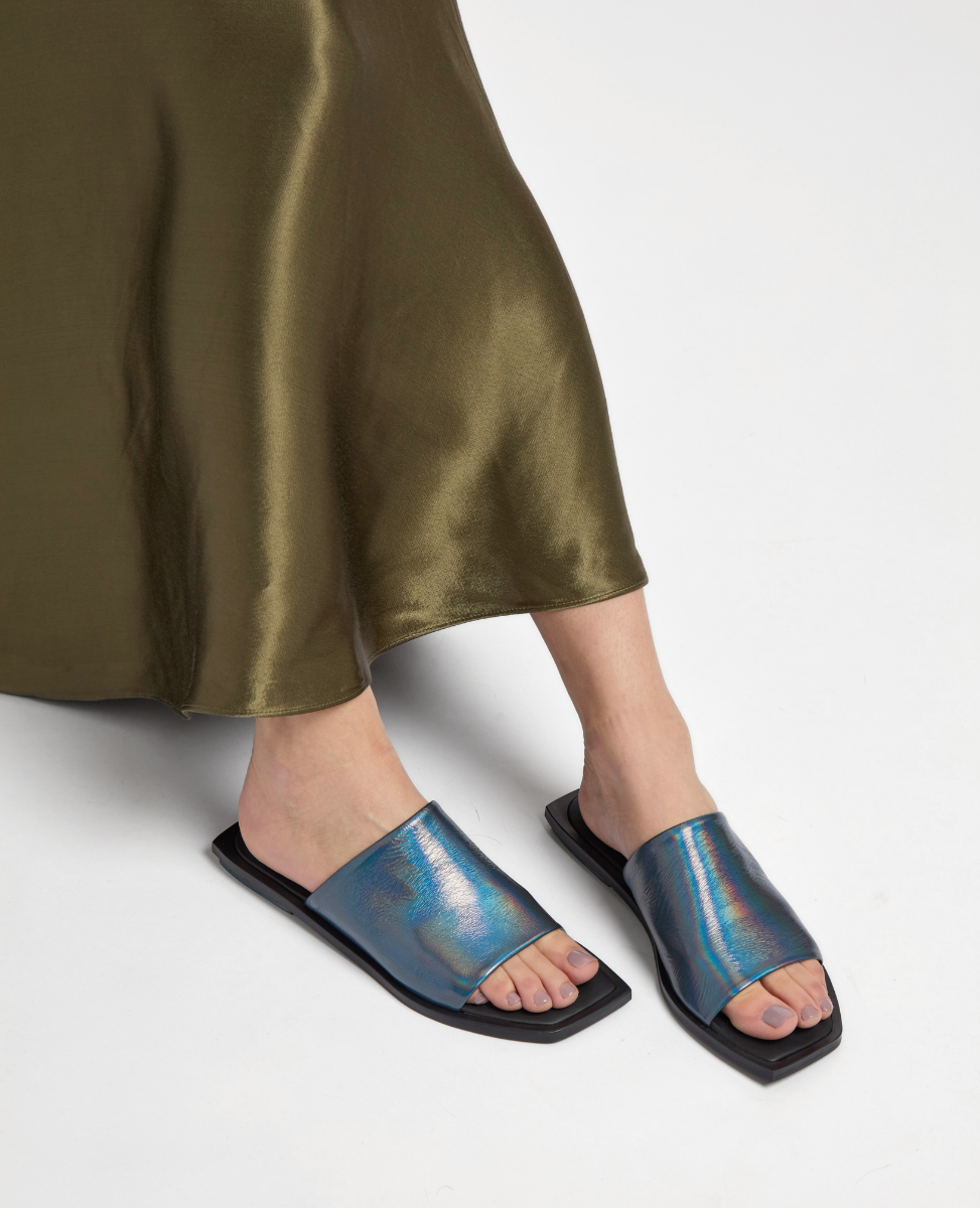A pair of iridescent blue slide sandals with a sleek, shiny finish, featuring wide straps and flat black soles, worn by a person in a flowing olive-green skirt.