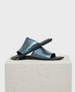 A pair of sleek, metallic blue slide sandals with a glossy finish, featuring a wide strap and flat black sole, displayed on a textured stone block. 1