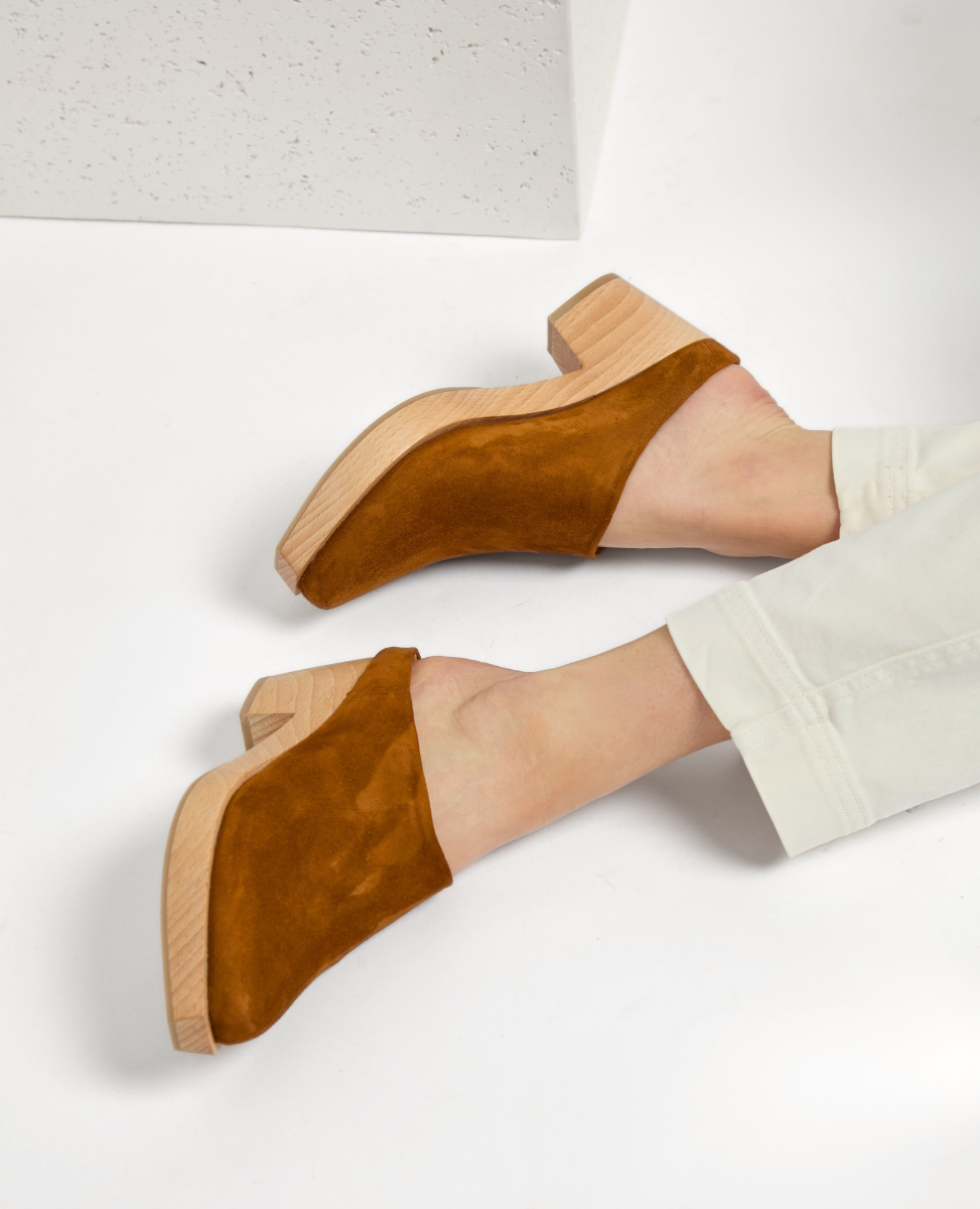 A pair of slip-on shoes with a rich brown suede upper, featuring a closed toe design and a chunky wooden heel, worn with light-colored pants.