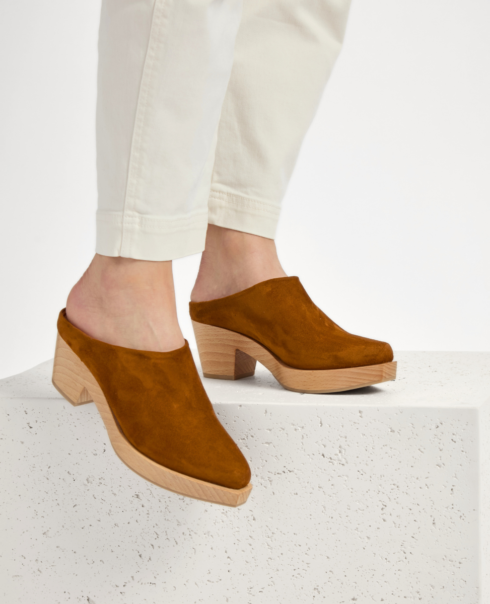 A pair of brown suede mules with a closed toe, featuring a chunky wooden heel and a smooth, minimalist design.