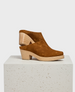 A pair of brown suede clogs with a smooth texture, featuring a closed toe design and a chunky, light wooden heel and sole, displayed on a textured white pedestal. 1