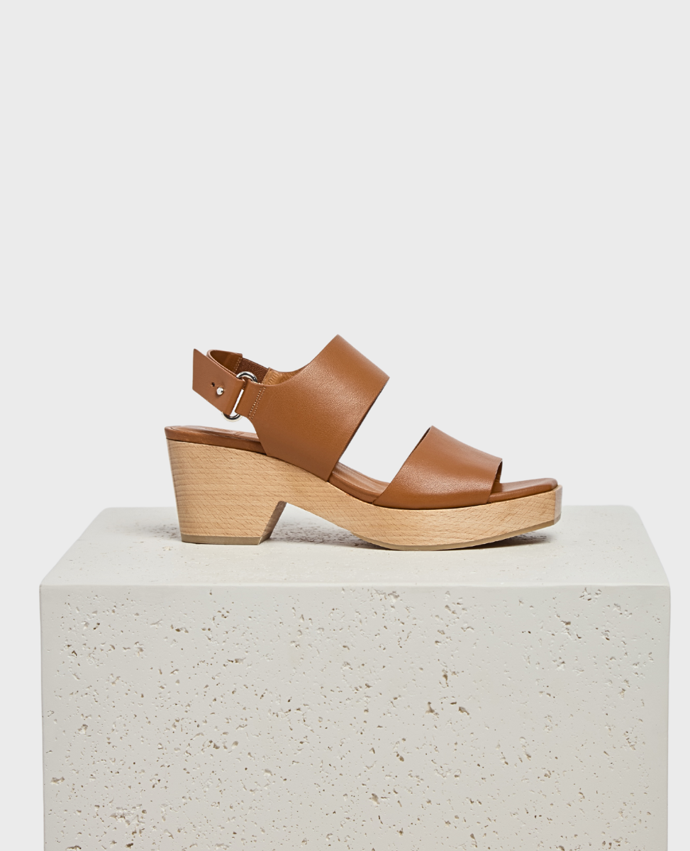 A brown leather sandal with two wide straps, an adjustable ankle buckle, and a wooden block heel, featuring a smooth texture and a minimalist design.