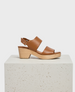 A brown leather sandal with two wide straps, an adjustable ankle buckle, and a wooden block heel, featuring a smooth texture and a minimalist design. 1